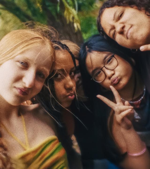 Ein Selfie zeigt Paula, Milli, Châo und Samira, die, ihre Köpfe aneinandergelehnt, gut gelaunt in die Kamera schauen. Châo und Samira machen ein Victory-Zeichen. Im Hintergrund sind Bäume und Wasser zu sehen.