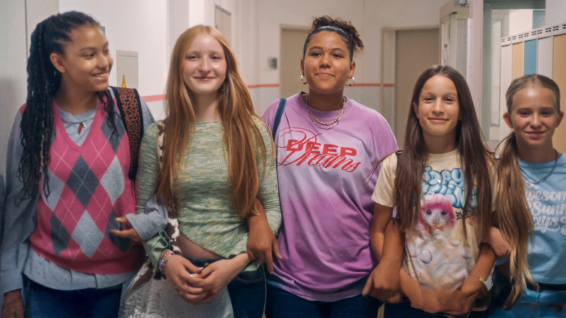 Milli, Paula, Samira, Liv und Sunny stehen freudestrahlend und eingehakt in die Kamera blickend nebeneinander im Schulflur. Nur Milli schaut an ihren Freundinnen entlang.