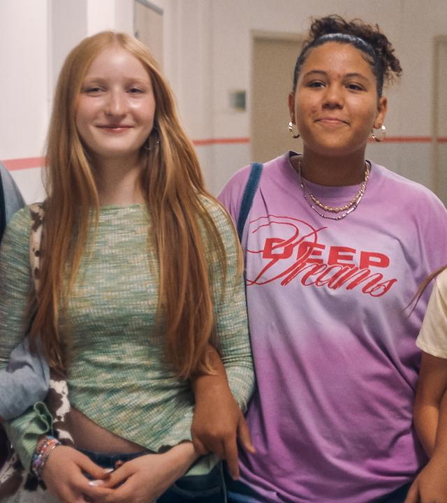 Milli, Paula, Samira, Liv und Sunny stehen freudestrahlend und eingehakt in die Kamera blickend nebeneinander im Schulflur. Nur Milli schaut an ihren Freundinnen entlang.