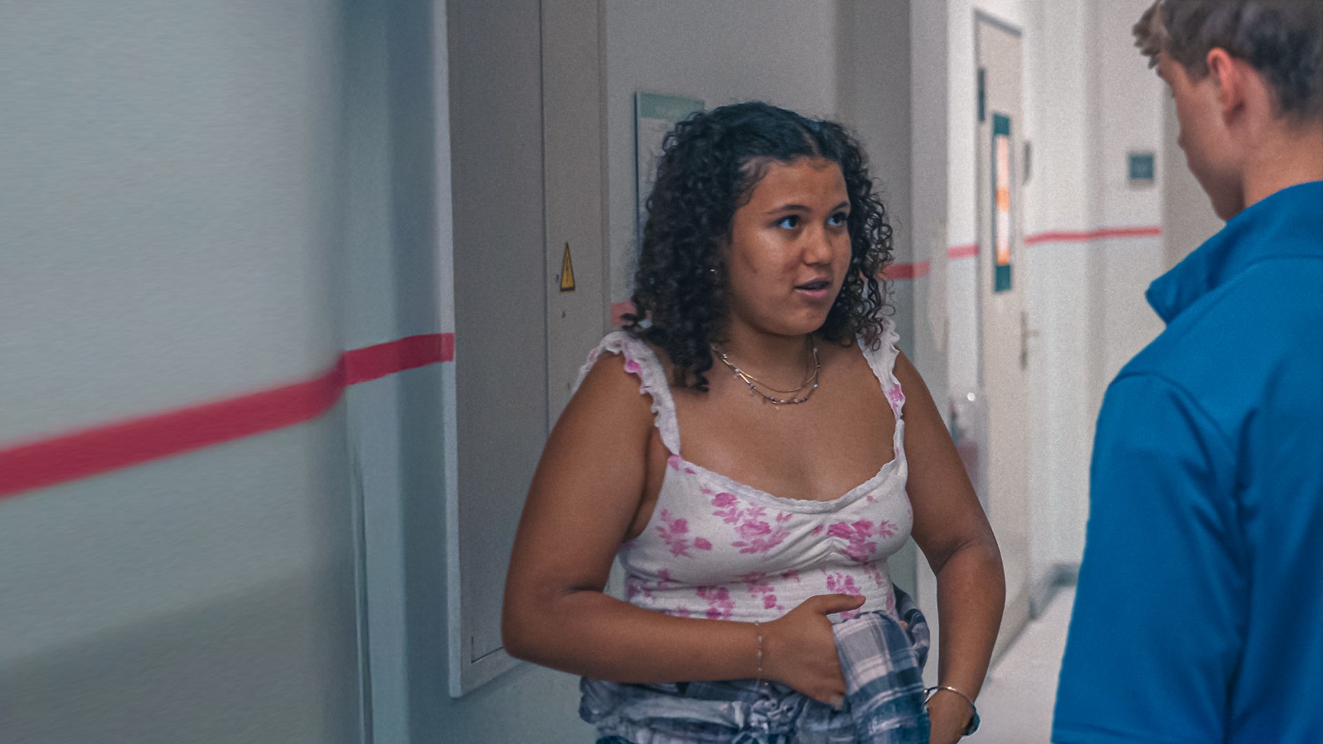 Samira spricht mit Lukas, der rechts von hinten zu sehen ist, auf dem Schulflur. Sie bindet sich ein kariertes Hemd um die Hüfte. Im Hintergrund sind die Türen zu den Klassenräumen zu sehen.