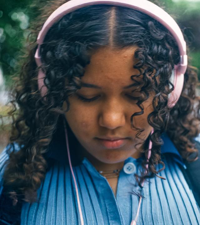 Samira ist mit nach unten geneigtem Kopf zu sehen. Sie trägt rosafarbene Kopfhörer und eine blaues Oberteil. Rechts von ihr ist ihr Handybildschirm eingeblendet, auf dem ein Video zum Thema Verliebtsein zu sehen ist.