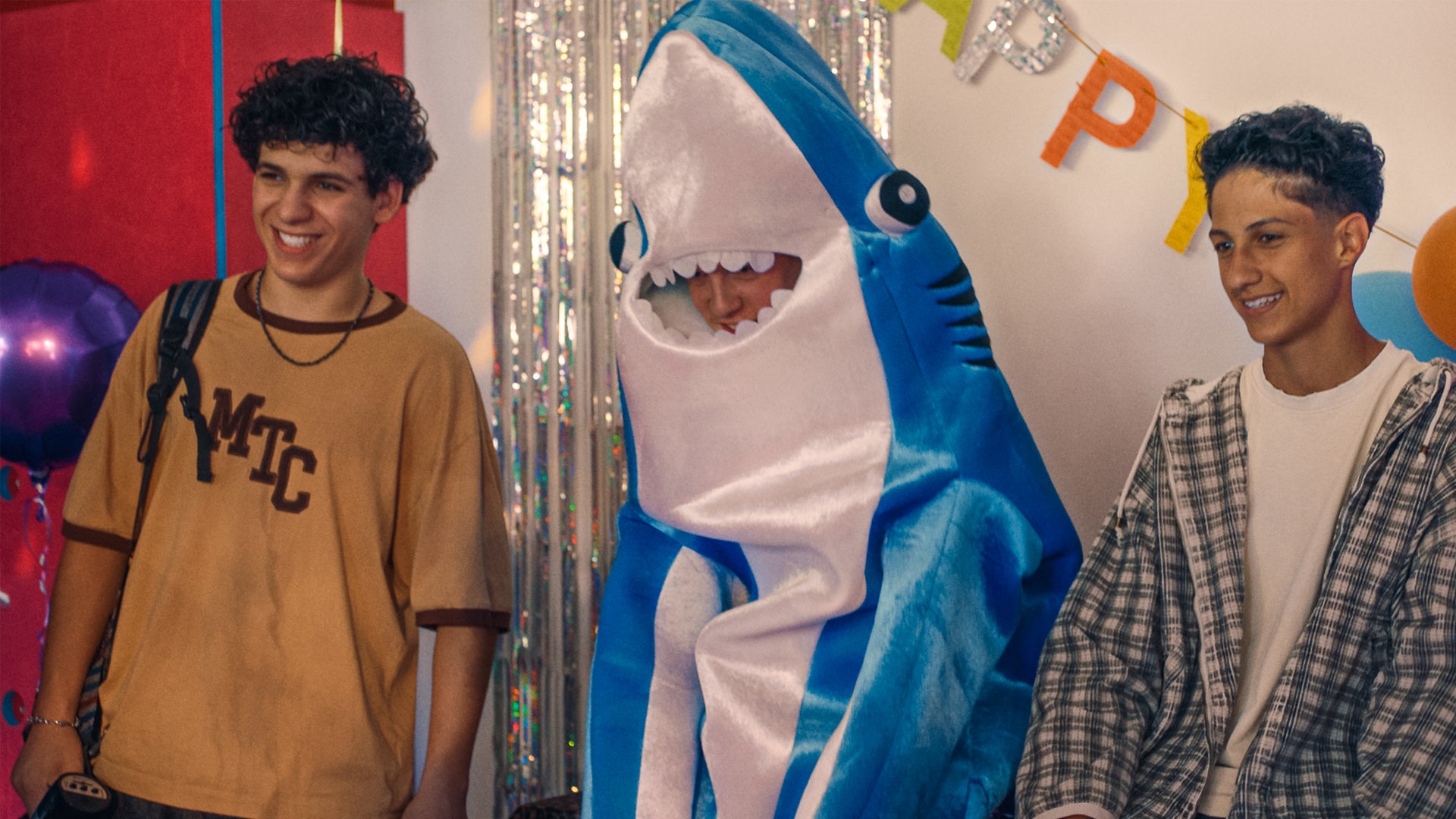 Enrico, Noah - der in einem Haifkostüm aus Plüsch steckt und nicht zu erkennen ist - und Pit stehen grinsend vor einer mit Lametta und einer Geburtstagsgirlande "Happy Birthday" geschmückten Wand im Boxstudio. Links schwebt ein lilafarbener Luftballon.