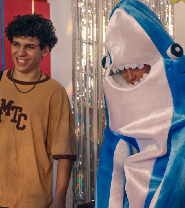 Enrico, Noah - der in einem Haifkostüm aus Plüsch steckt und nicht zu erkennen ist - und Pit stehen grinsend vor einer mit Lametta und einer Geburtstagsgirlande "Happy Birthday" geschmückten Wand im Boxstudio. Links schwebt ein lilafarbener Luftballon.