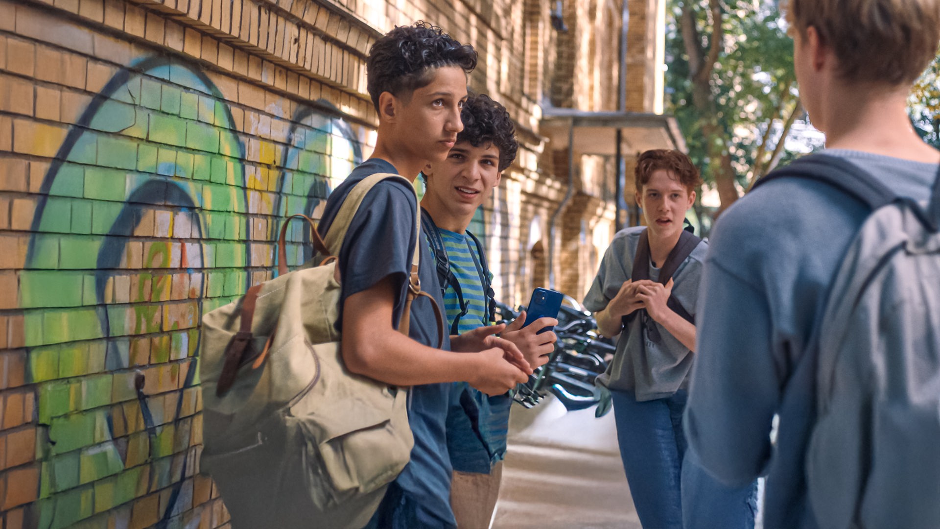 Pit steht mit seinem Schulrucksack über der Schulter auf dem Pausenhof mit Enrico und Noah und blickt Lukas, der von hinten zu sehen ist und auch einen Schulrucksack trägt, an. Enrico hat ein Handy in der Hand und schaut zu Pit, Noah sieht Lukas an, der gerade spricht. Links ist die Backsteinwand der Schule mit Grafitti zu sehen.