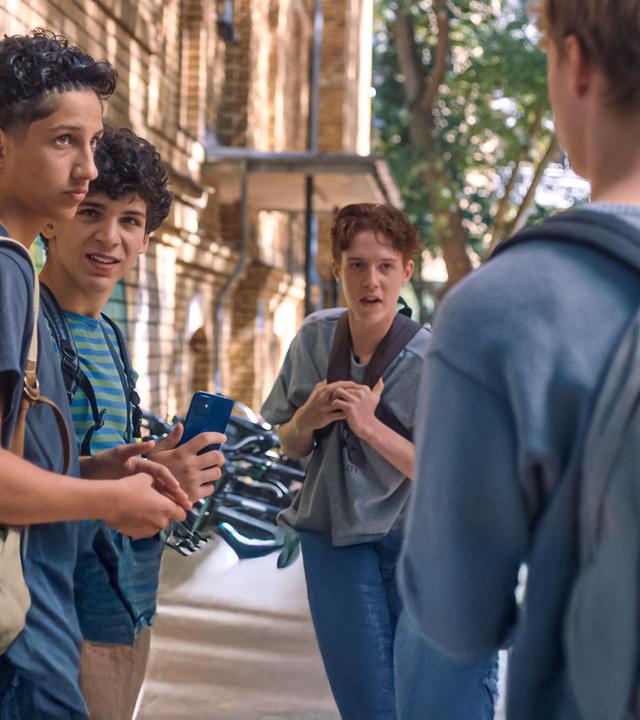Pit steht mit seinem Schulrucksack über der Schulter auf dem Pausenhof mit Enrico und Noah und blickt Lukas, der von hinten zu sehen ist und auch einen Schulrucksack trägt, an. Enrico hat ein Handy in der Hand und schaut zu Pit, Noah sieht Lukas an, der gerade spricht. Links ist die Backsteinwand der Schule mit Grafitti zu sehen.