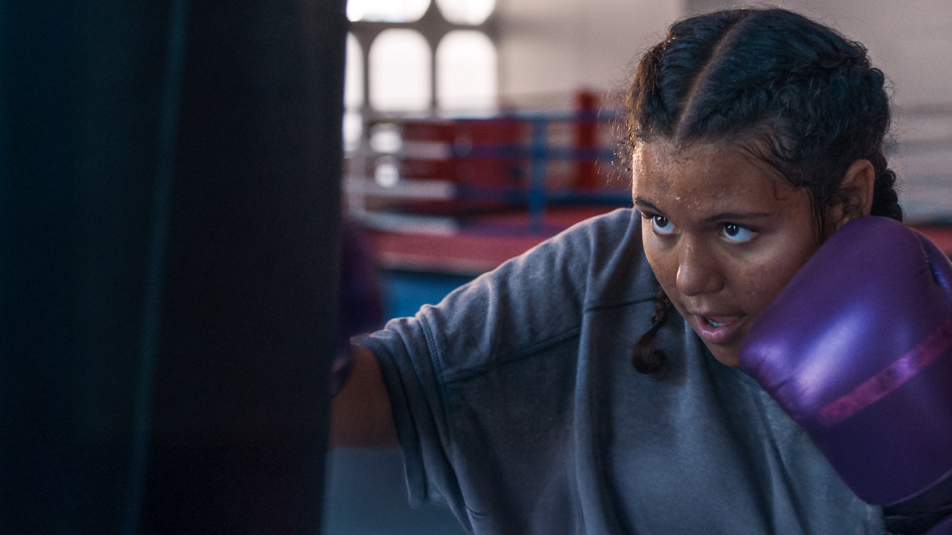 Samira ist im Boxstudio und boxt hoch konzentriert und leicht verschwitzt mit lilafarbenen Boxhandschuhen auf einen Boxsack ein. Ihr Haare sind zu zwei Zöpfen nach hinten geflochten.