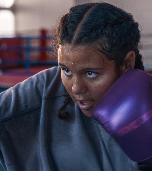 Samira ist im Boxstudio und boxt hoch konzentriert und leicht verschwitzt mit lilafarbenen Boxhandschuhen auf einen Boxsack ein. Ihr Haare sind zu zwei Zöpfen nach hinten geflochten.