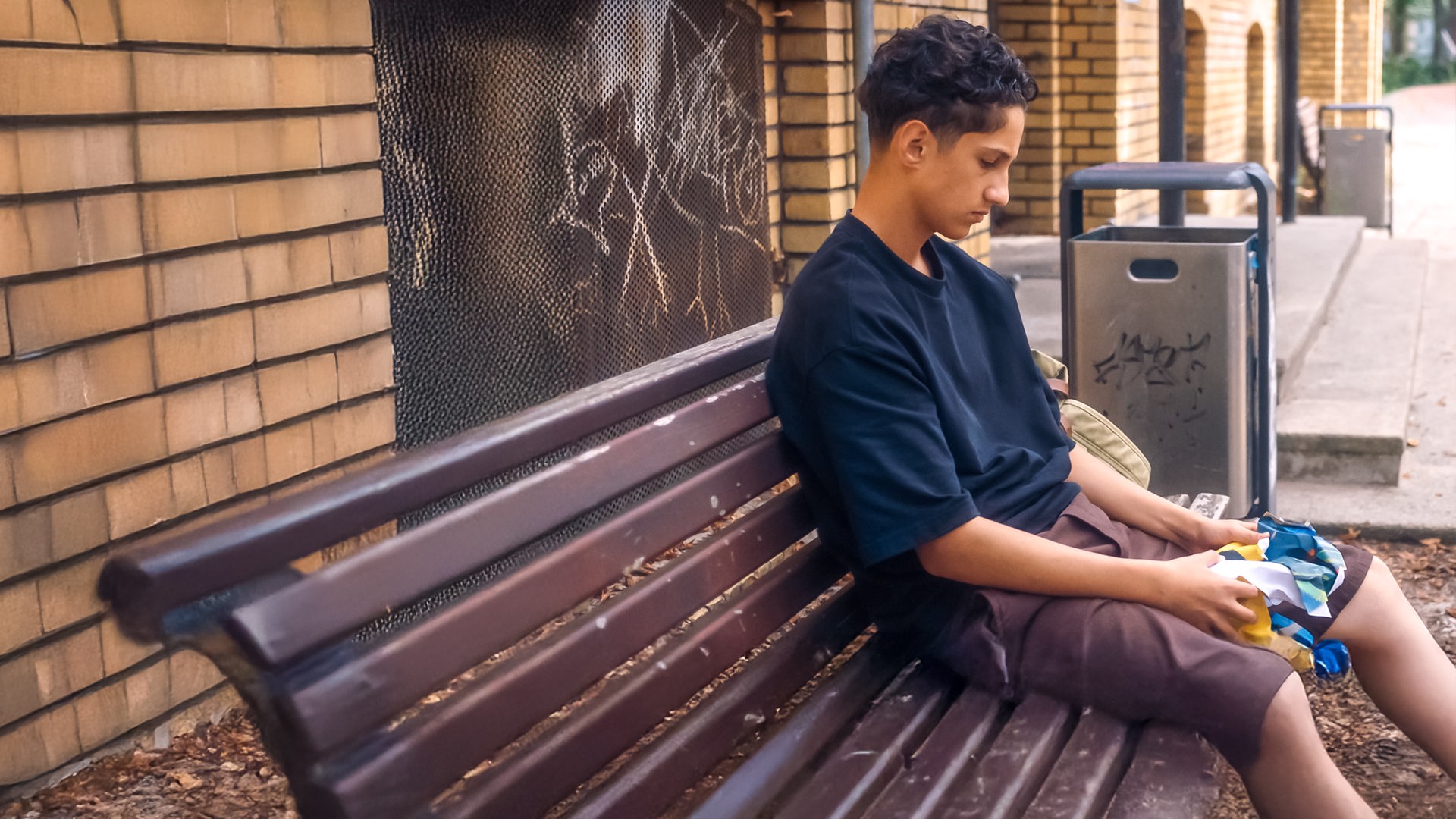 Pit sitzt niedergeschlagen auf einer braunen Holzbank. Er hält ein buntes Tuch in den Händen und hat den Blick nach unten gerichtet auf seine Schuhe. Links ist ein helles Backsteingebäude zu sehen mit Grafitti an einem Gitter. Zwischen zwei Mülleimern führt eine Treppe in das Backsteingebäude.