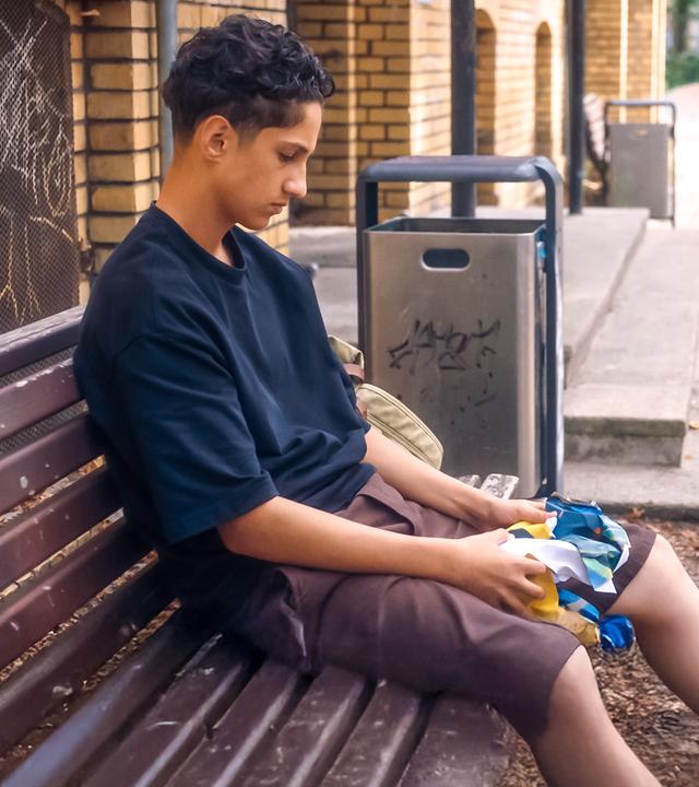 Pit sitzt niedergeschlagen auf einer braunen Holzbank. Er hält ein buntes Tuch in den Händen und hat den Blick nach unten gerichtet auf seine Schuhe. Links ist ein helles Backsteingebäude zu sehen mit Grafitti an einem Gitter. Zwischen zwei Mülleimern führt eine Treppe in das Backsteingebäude.