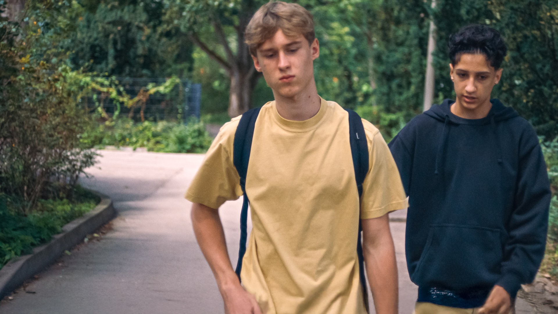 Lukas läuft mit gesenktem Blick und niedergeschlagen eine Straße entlang. Er hat einen Schulrucksack auf, Pit läuft leicht hinter ihm in einem dunkelblauen Sweatshirt.
