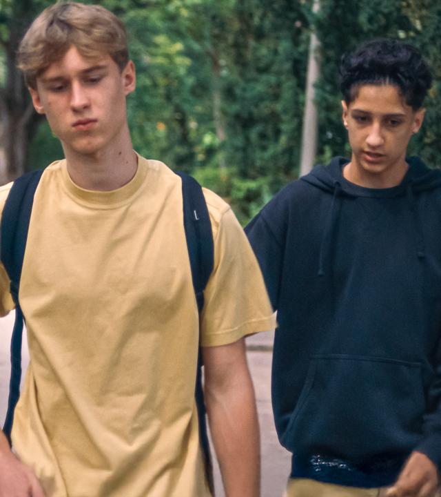 Lukas läuft mit gesenktem Blick und niedergeschlagen eine Straße entlang. Er hat einen Schulrucksack auf, Pit läuft leicht hinter ihm in einem dunkelblauen Sweatshirt.