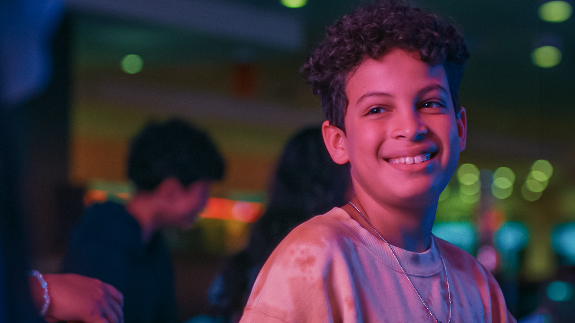 Tom steht strahlend im Bowlingcenter, sein Gesicht ist in rötliches Licht getaucht. Im Hintergrund ist Pit zu erkennen und andere Kinder. Blaue, grüne und pinke Spots sind rechts zu sehen.
