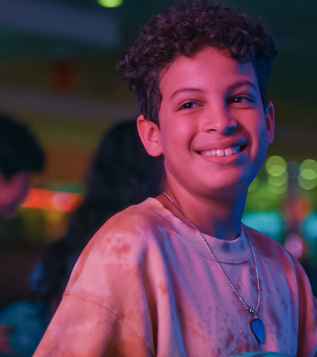 Tom steht strahlend im Bowlingcenter, sein Gesicht ist in rötliches Licht getaucht. Im Hintergrund ist Pit zu erkennen und andere Kinder. Blaue, grüne und pinke Spots sind rechts zu sehen.