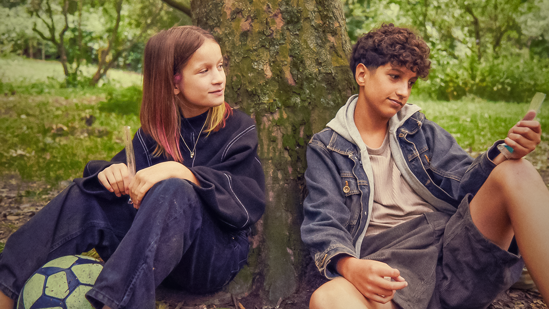 Liv und Pit sitzen im Grünen unter einem Baum. Beide haben ein Eis in einer schmalen durchsichtigen Tüte in der Hand. Vor Liv liegt ein Fußball. Liv blickt lächelnd und ein bisschen erwartungsvoll zu Pit, der leicht lächelnd nach vorne auf sein Eis schaut.