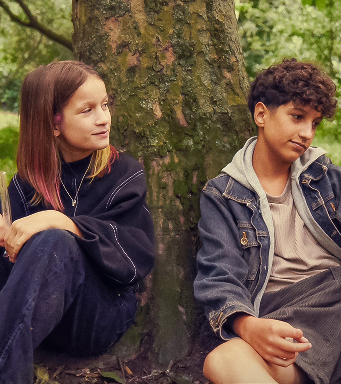 Liv und Pit sitzen im Grünen unter einem Baum. Beide haben ein Eis in einer schmalen durchsichtigen Tüte in der Hand. Vor Liv liegt ein Fußball. Liv blickt lächelnd und ein bisschen erwartungsvoll zu Pit, der leicht lächelnd nach vorne auf sein Eis schaut.