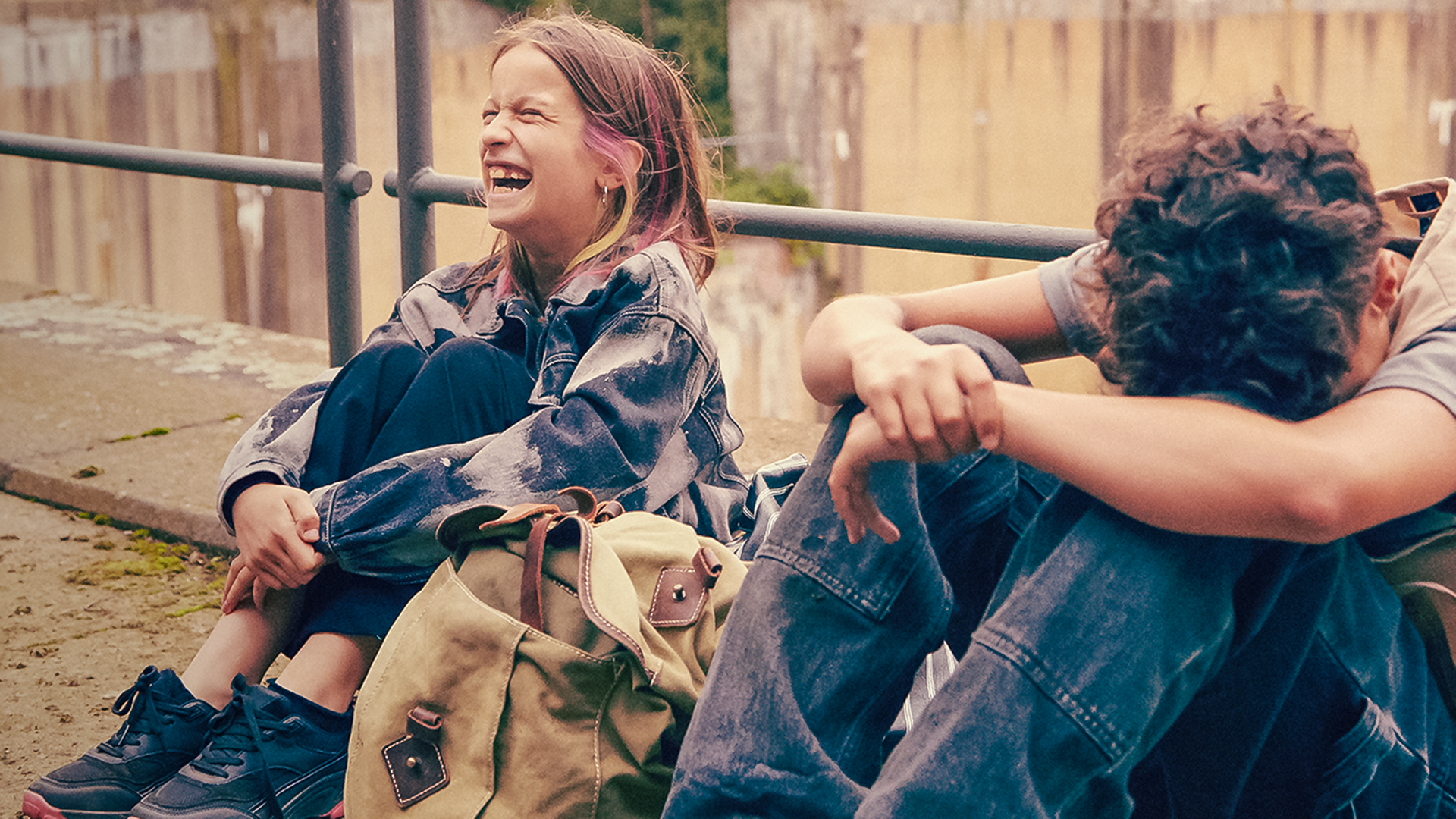  Am Rand eines stillgelegten Gasometers sitzen Liv und Pit am Geländer. Zwischen ihnen steht Pits Rucksack. Liv lacht lauthals, Pit hat seinen Kopf auf seine Knie gelegt, die er mit verschränkten Armen umfasst.