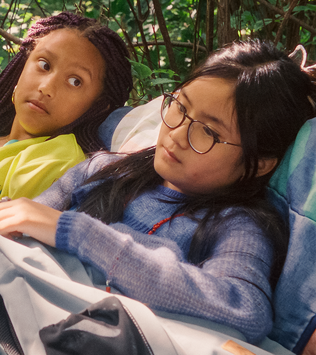 Milli und Châu liegen mit leicht aufgerichtetem Oberkörper unter einem grünen Busch auf einem Lager aus Decken. Châu blickt traurig in die Ferne, während Milli leicht fragend zu ihr herüberschaut. Die Sonne scheint und sie haben ihre Rucksäcke bei sich.