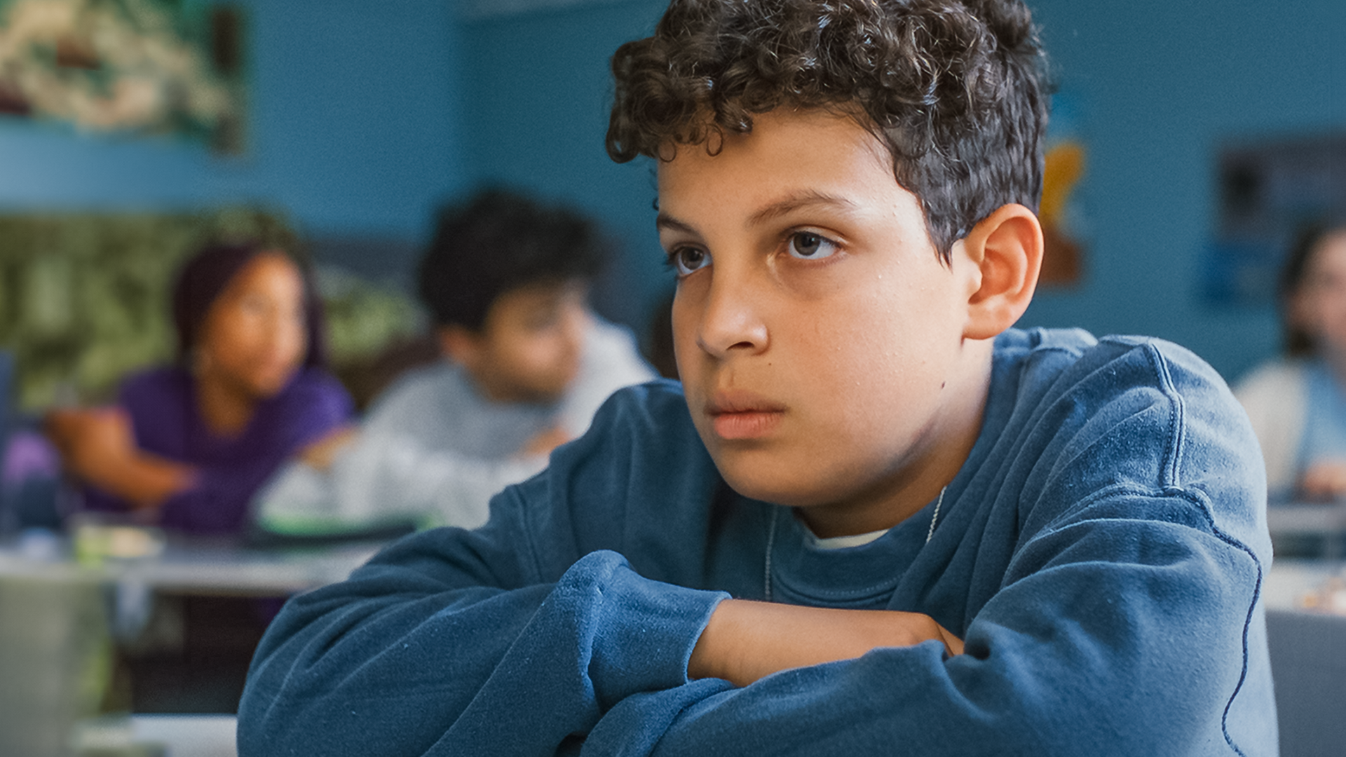  Im Klassenraum sitzt Tom mit verschränkten Armen an seinem Tisch und schaut traurig nach vorne. Im Hintergrund sind noch andere Mitschüler verschwommen zu erkennen.