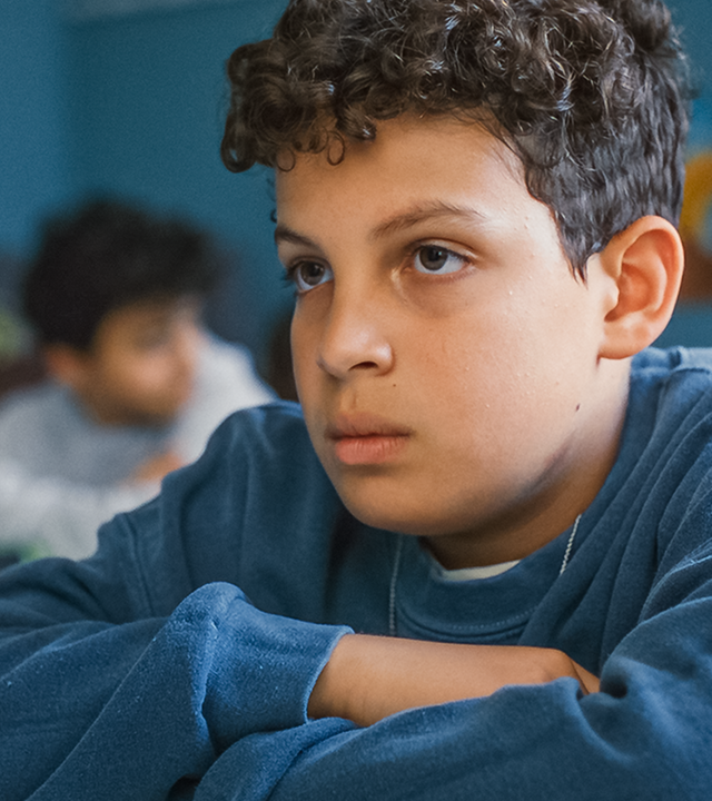  Im Klassenraum sitzt Tom mit verschränkten Armen an seinem Tisch und schaut traurig nach vorne. Im Hintergrund sind noch andere Mitschüler verschwommen zu erkennen.