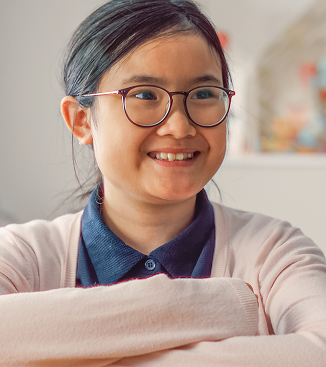 Châu sitzt grinsend mit verschränkten Armen bei ihren Eltern in der Küche und sieht glücklich aus. Sie trägt eine dunkelblaue Bluse und eine hellrosa Strickjacke.