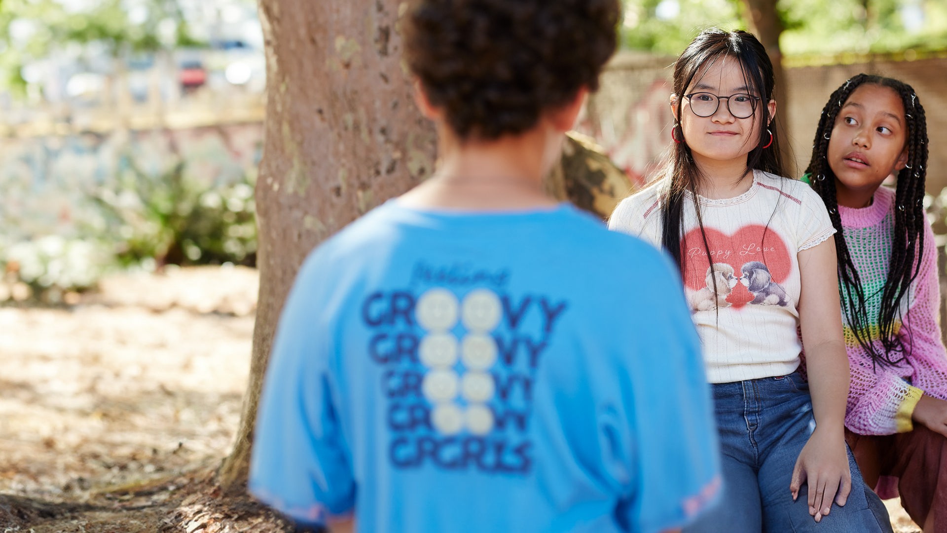 Draußen bei Sonnenlicht auf dem Schulhof steht Tom vor Châu und Milli. Châu, die ihm gegenüber steht, schaut mit einem verkniffenen Lächeln zu Tom, während Milli, die seitlich hinter Châu sitzt, neugierig zu ihrer Freundin guckt.