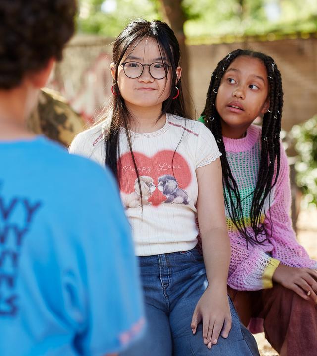 Draußen bei Sonnenlicht auf dem Schulhof steht Tom vor Châu und Milli. Châu, die ihm gegenüber steht, schaut mit einem verkniffenen Lächeln zu Tom, während Milli, die seitlich hinter Châu sitzt, neugierig zu ihrer Freundin guckt.