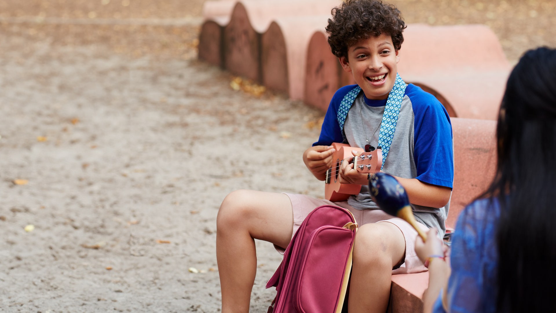 Auf dem Schulhof sitzen sich Tom und Châu gegenüber. Tom spielt seine Ukulele und schaut Châu lächelnd an. Diese sitzt mit dem Rücken zum Betrachter und hält Maracas in den Händen.