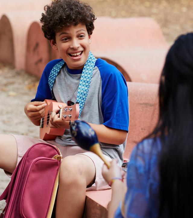 Auf dem Schulhof sitzen sich Tom und Châu gegenüber. Tom spielt seine Ukulele und schaut Châu lächelnd an. Diese sitzt mit dem Rücken zum Betrachter und hält Maracas in den Händen.