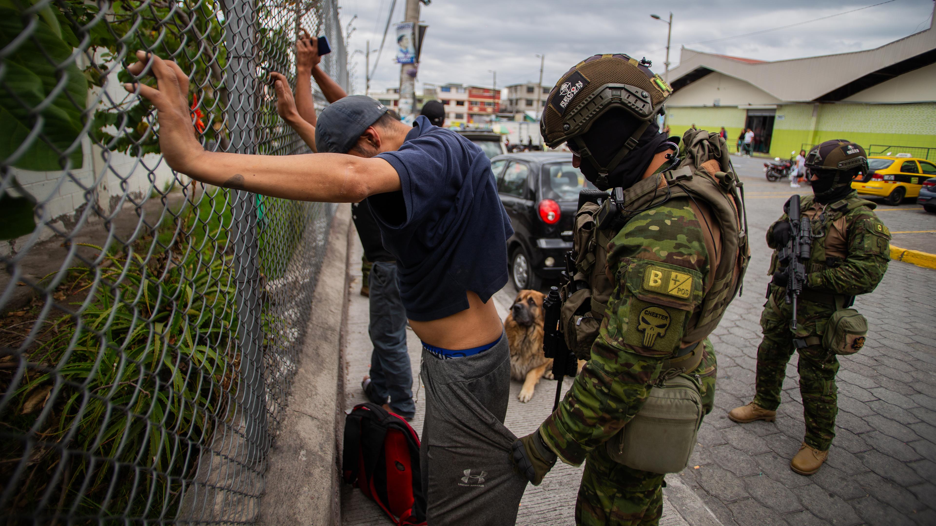 Ein Soldat kontrolliert die Taschen eines mit dem Rücken zu ihm stehenden Mannes, der seine Hände an einen Zaun gelegt hat.