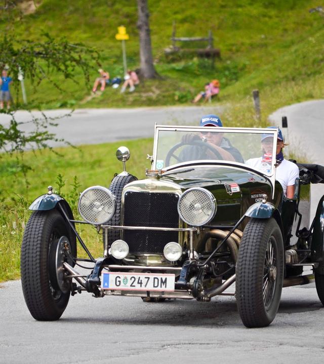 Ein dunkelgrüner Sunbeam Supersport Oldtimer mit zwei Insassen fährt eine Bergstraße entlang, im Hintergrund Zuschauer auf einer Wiese.