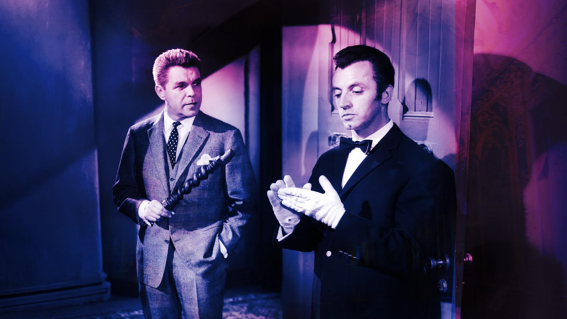 Ein Mann im Anzug hält ein hölzernes Objekt und schaut einen Butler in Livree an, der sich weiße Handschuhe anzieht.