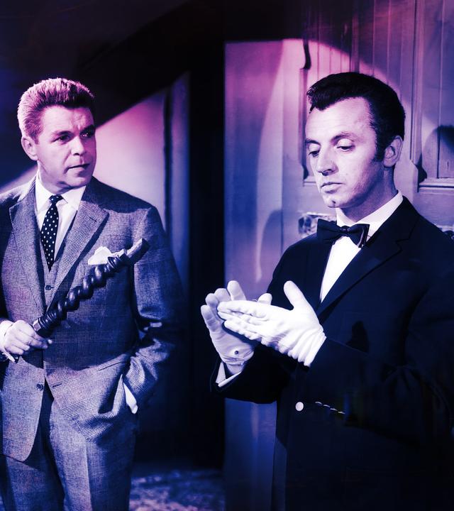 Ein Mann im Anzug hält ein hölzernes Objekt und schaut einen Butler in Livree an, der sich weiße Handschuhe anzieht.