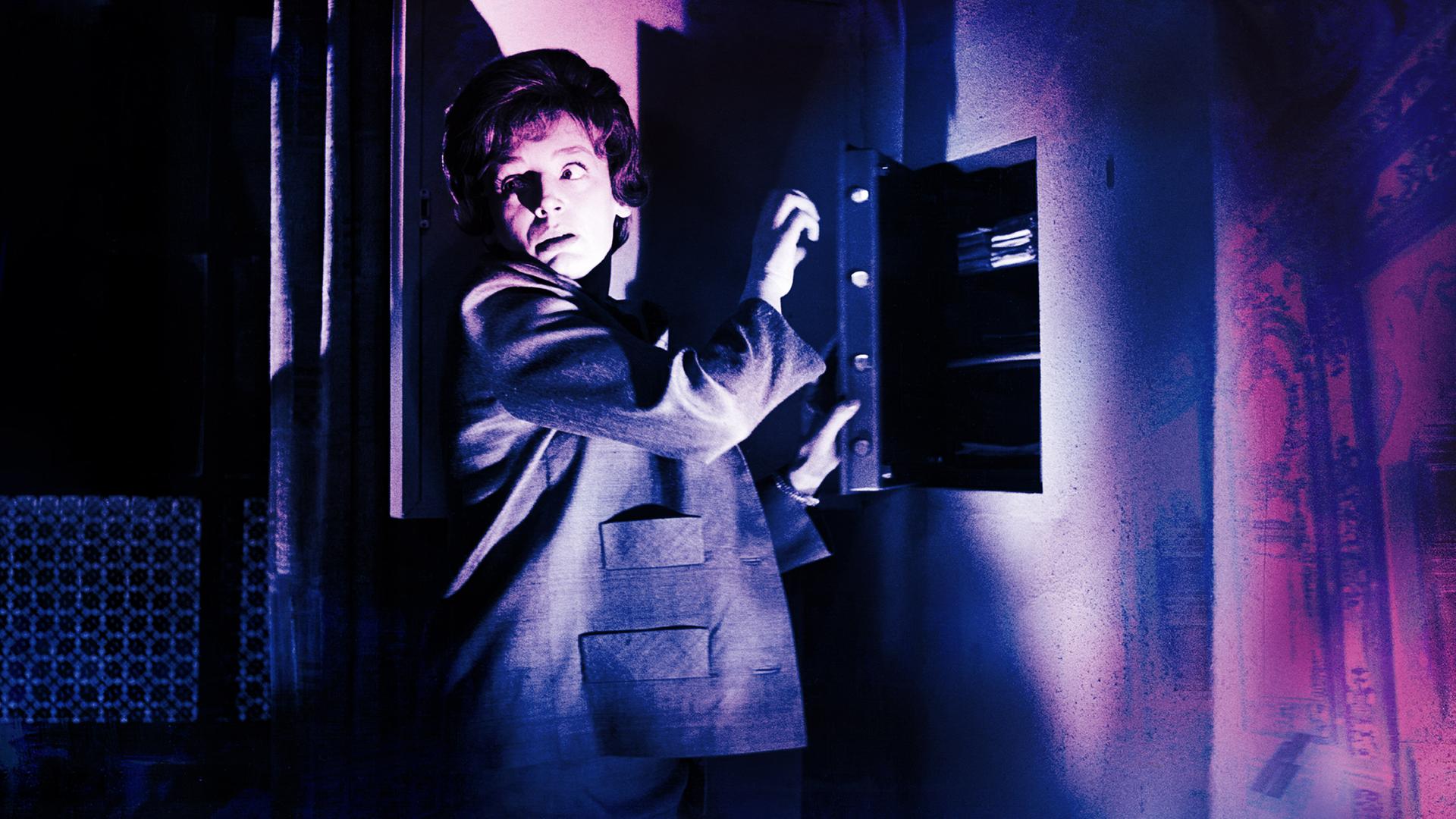 Eine Frau steht vor einem halb geöffneten Wandsafe im Halbschatten, schlaglichtartig wird ihr Gesicht von einer einzelnen Lichtquelle erhellt. Vorsichtig blickt sie über ihre Schulter.