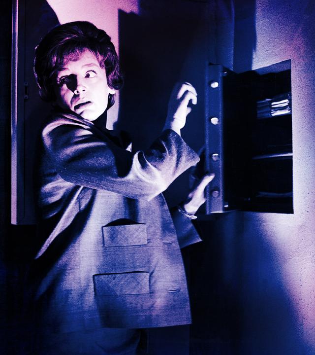 Eine Frau steht vor einem halb geöffneten Wandsafe im Halbschatten, schlaglichtartig wird ihr Gesicht von einer einzelnen Lichtquelle erhellt. Vorsichtig blickt sie über ihre Schulter.