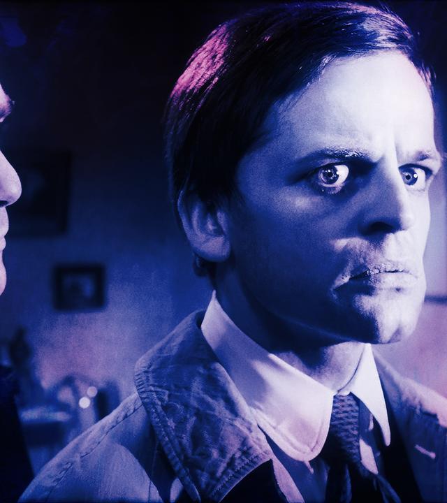 Zwei Männer in einem Raum  einer blickt kritisch auf den anderen, der mit erschrockenem Gesichtsausdruck zur Seite schaut.