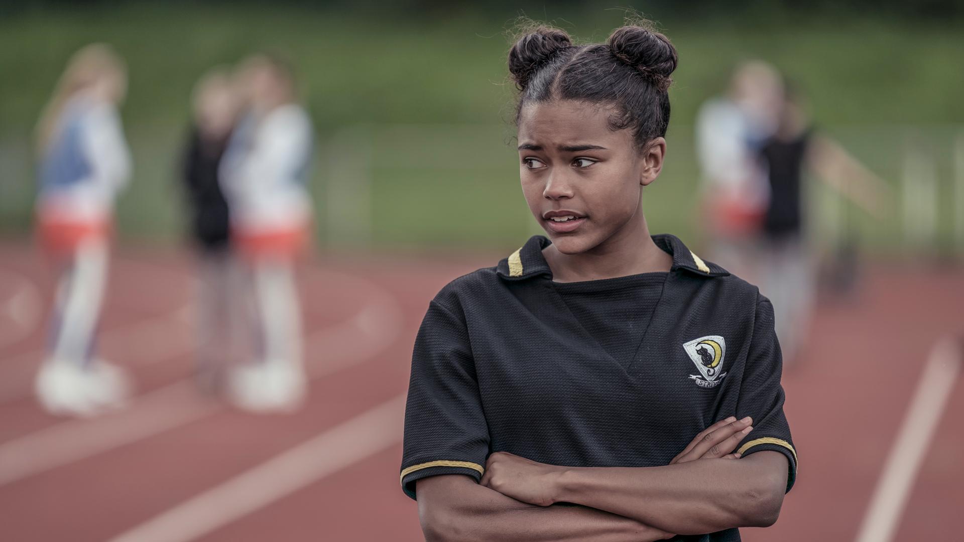 Auf dem Sportplatz steht Edith, sie hält die Arme verschränkt vor sich und blickt grüblerisch nach unten.