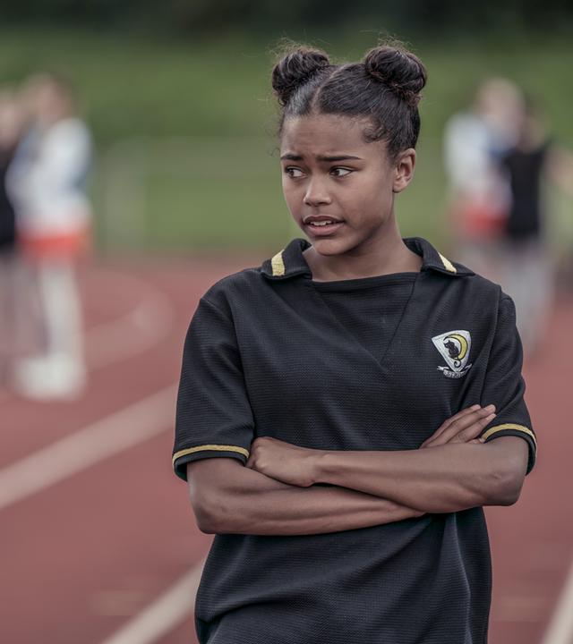 Auf dem Sportplatz steht Edith, sie hält die Arme verschränkt vor sich und blickt grüblerisch nach unten.