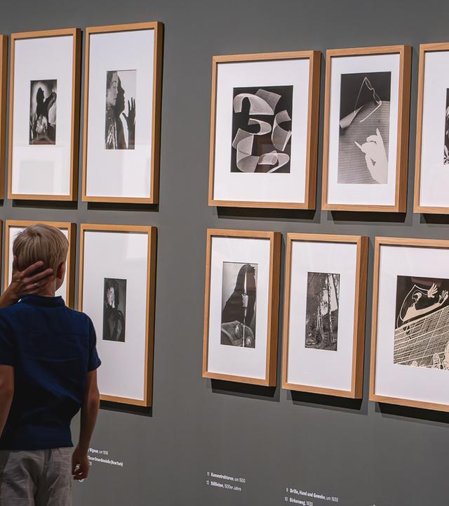 Eine Frau und ein Kind schauen sich Schwarz-Weiß-Fotos und Fotogramme an, die in einem Ausstellungsraum an der Wand hängen.