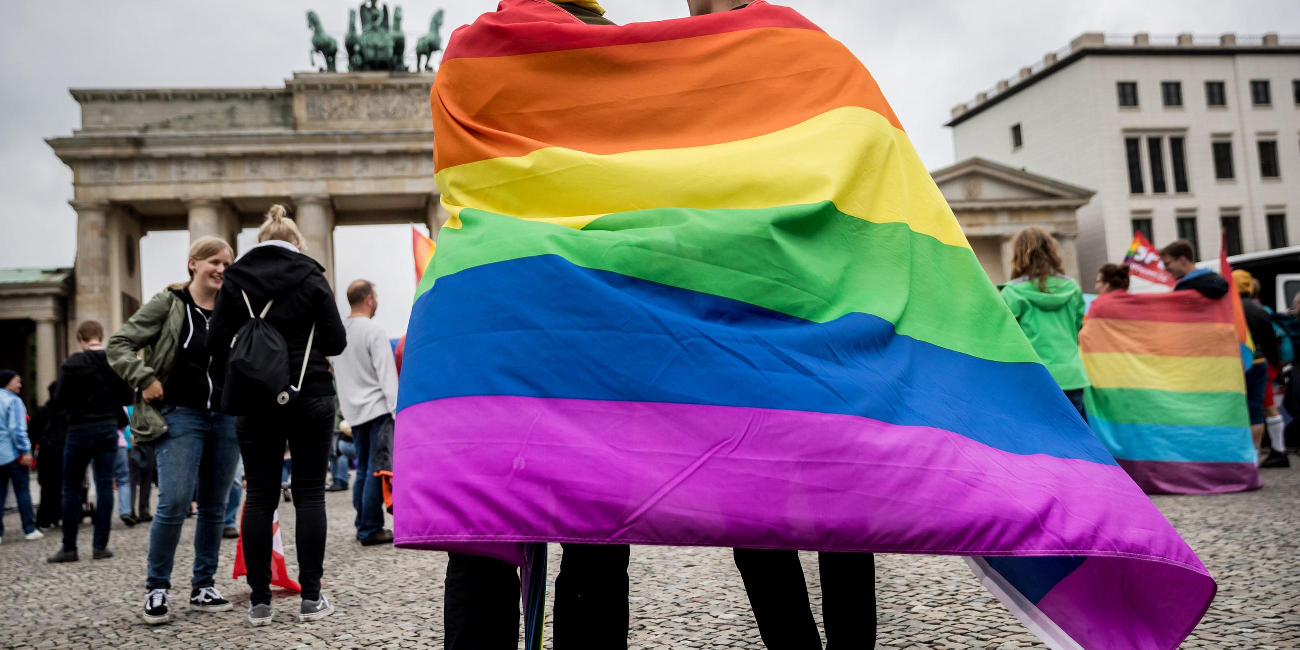 Zwei Männer stehen vor dem Brandenburger Tor in Berlin mit einer Regenbogenflagge.