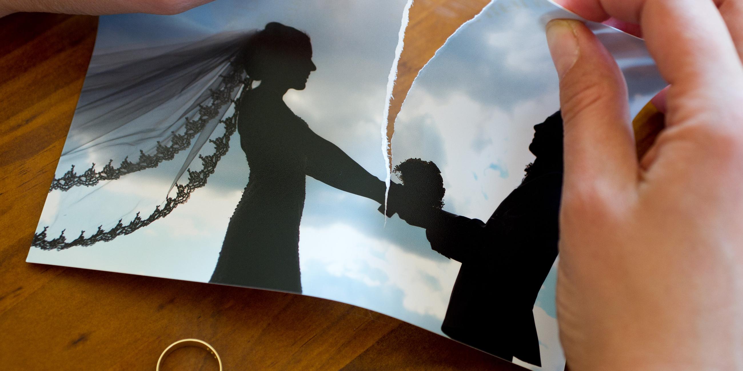Hände zereißen ein Hochzeitsfoto, unter dem Foto liegt ein Ehering.