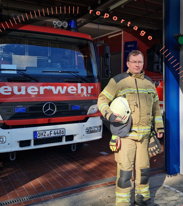 Chris Hartmann steht vor einer Garage mit Einsatzfahrzeugen der Feuerwehr. Er schaut in die Kamera.
