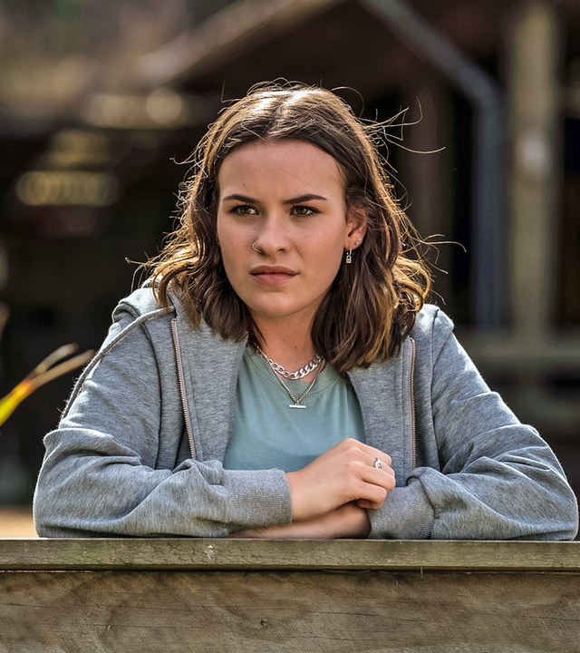 Issie steht argwöhnisch blickend am Rande der Pferdekoppel und stützt ihre Arme auf dem Gatter auf.