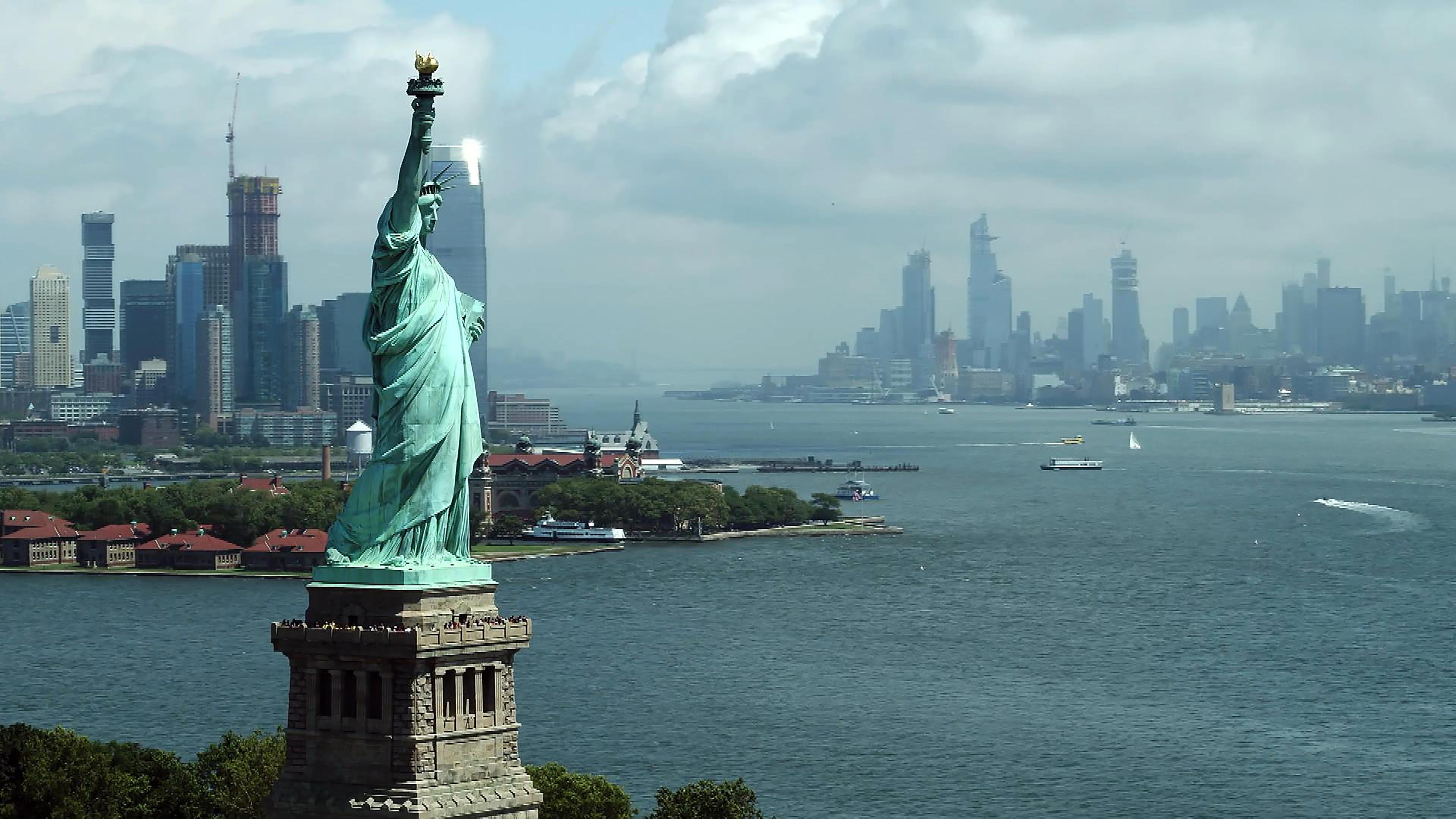 "Eiffels Superbauten - Giganten aus Stahl und Eisen": Vorne links, die Freiheitstatue von New York City, Wasser mit Booten, im Hintergrund die Skyline der Stadt.