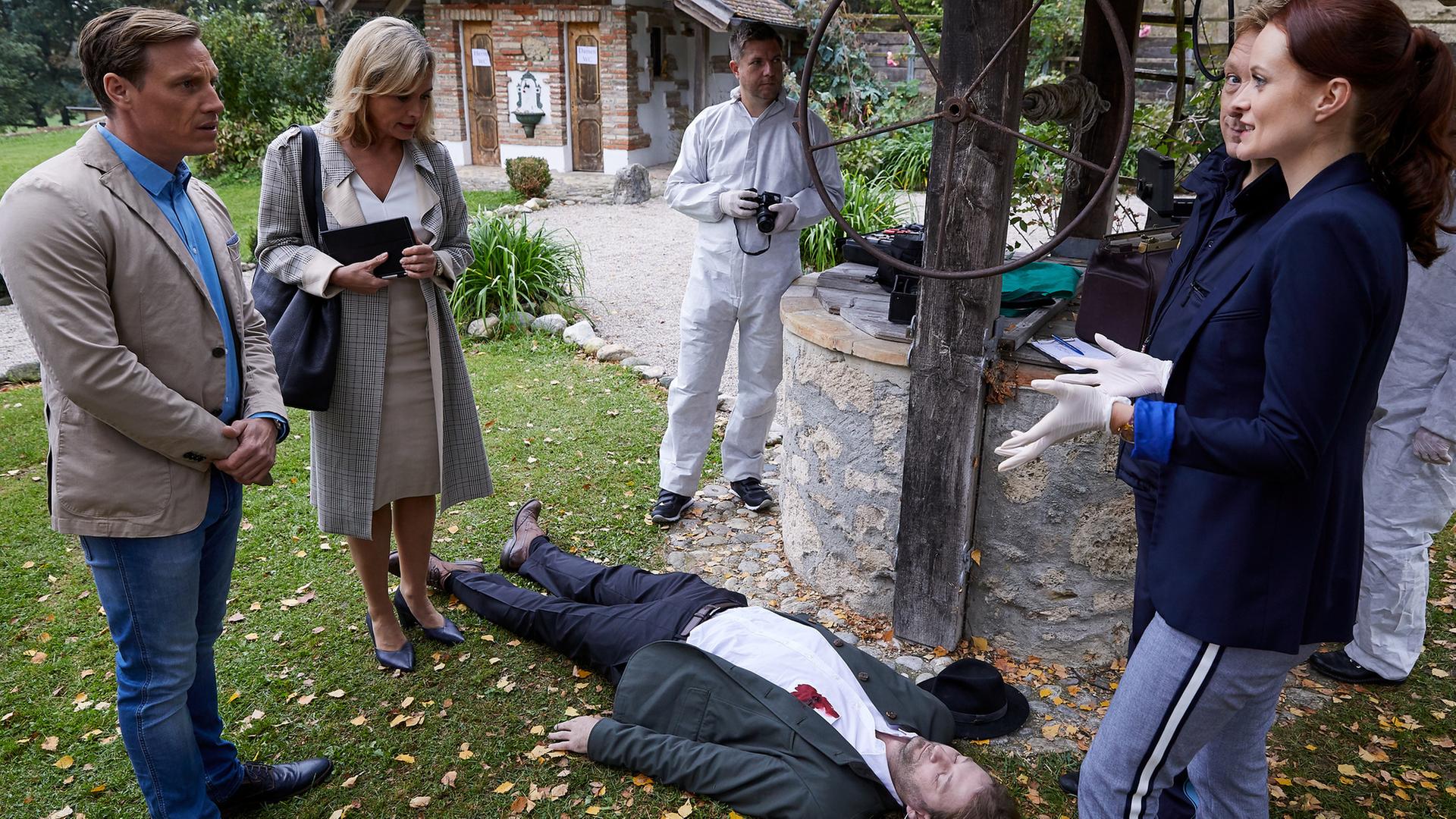 "Die Rosenheim-Cops - Ein Auftrag für Frau Stockl": Pathologin Mai (Sina Wilke) steht mit Latexhandschuhen vor dem Mordopfer (Komparse), bei dem sich ein Blutfleck in Brusthöhe auf dem weißen Hemd abzeichnet. Um das Opfer herum stehen die Kommissare Bach (Patrick Kalupa) und Danner (Katharina Abt) sowie zwei Kollegen der KTU (Komparsen) in weißer Schutzkleidung. Neben ihnen ist ein alter Brunnen mit Förderrad zu sehen, im Hintergrund ein kleines altes Haus.
