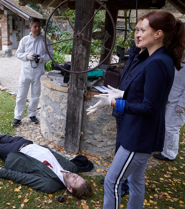 "Die Rosenheim-Cops - Ein Auftrag für Frau Stockl": Pathologin Mai (Sina Wilke) steht mit Latexhandschuhen vor dem Mordopfer (Komparse), bei dem sich ein Blutfleck in Brusthöhe auf dem weißen Hemd abzeichnet. Um das Opfer herum stehen die Kommissare Bach (Patrick Kalupa) und Danner (Katharina Abt) sowie zwei Kollegen der KTU (Komparsen) in weißer Schutzkleidung. Neben ihnen ist ein alter Brunnen mit Förderrad zu sehen, im Hintergrund ein kleines altes Haus.