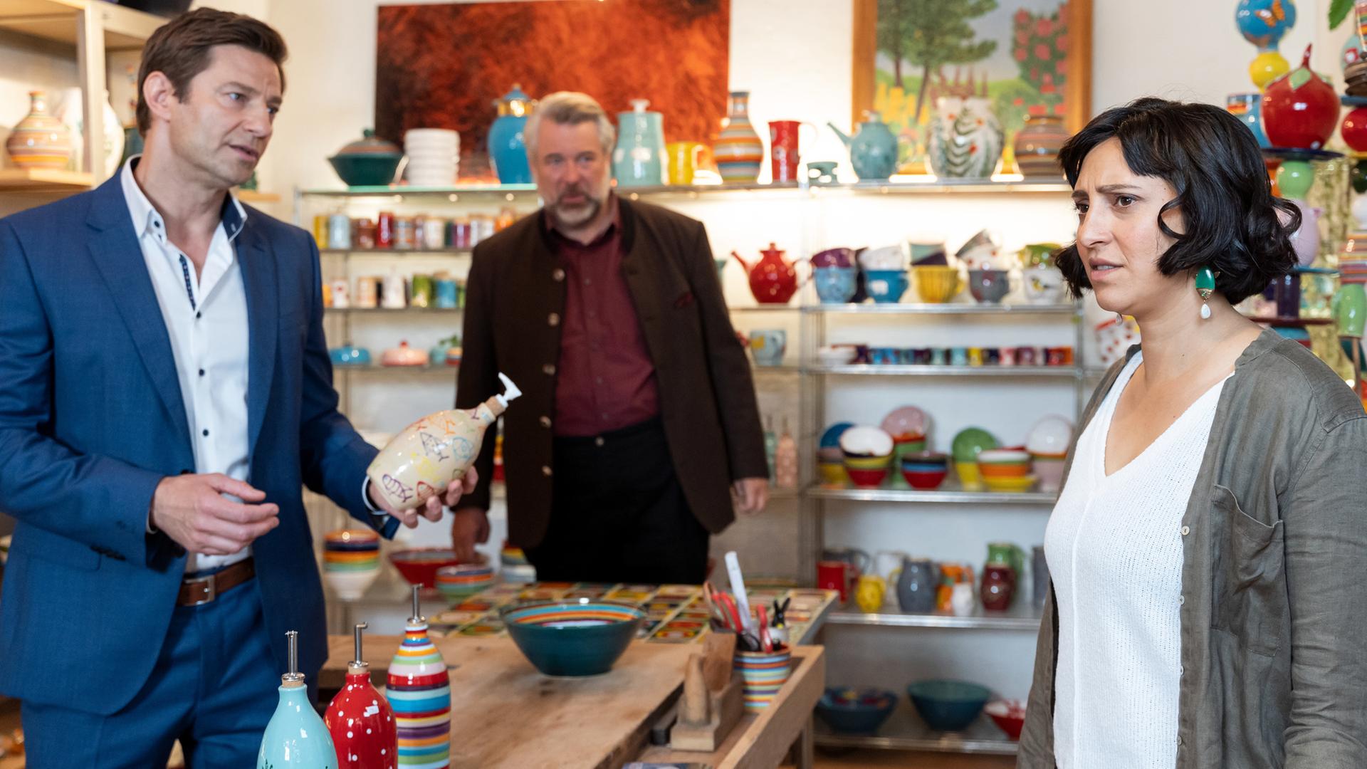 "Die Rosenheim-Cops - Ein Date für Stadler": Die Kommissare Hansen (Igor Jeftić) und Stadler (Dieter Fischer) stehen mit Franziska Köhler (Mira Mazumdar) in deren Keramikgeschäft und unterhalten sich. Hansen hält eine Keramikflasche in den Händen. Auf dem Tisch im Vordergrund sowie in den Regalen im Hintergrund sind zahlreiche bunte Keramikartikel zu sehen.