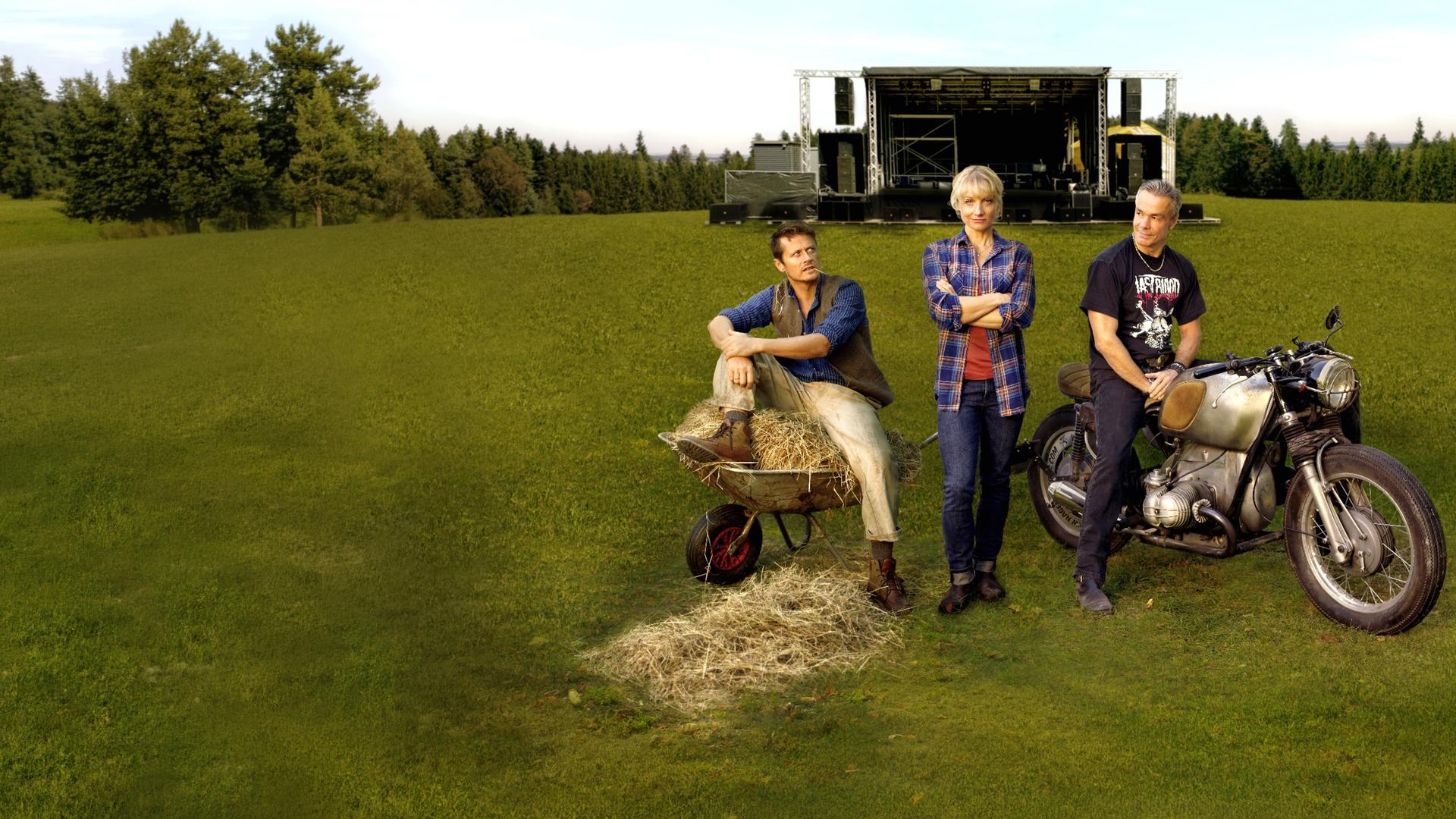  Paul (Roman Knizka), Caro (Christina Große) und Mike (Hannes Jaenicke) stehen auf einer Wiese. Weit im Hintergrund ist die große Bühne zu sehen.