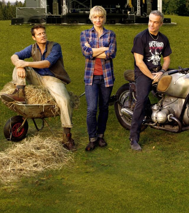  Paul (Roman Knizka), Caro (Christina Große) und Mike (Hannes Jaenicke) stehen auf einer Wiese. Weit im Hintergrund ist die große Bühne zu sehen.