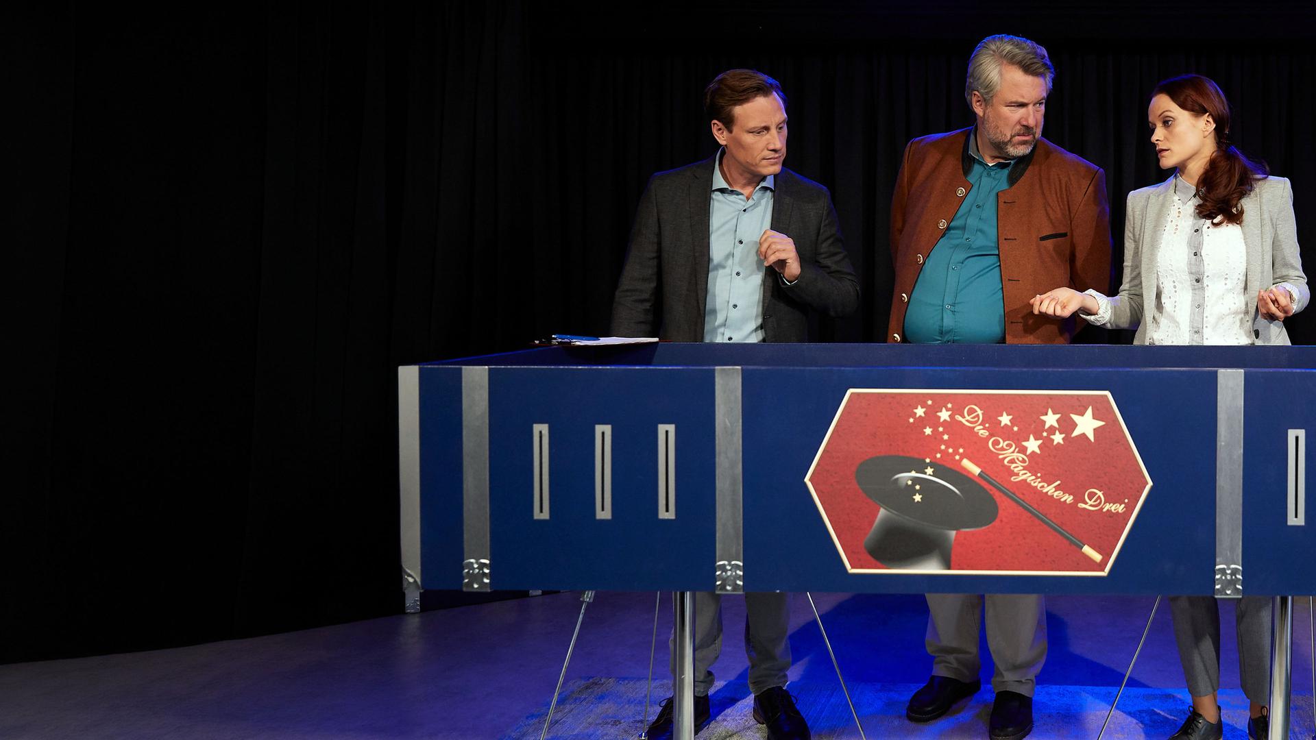 "Die Rosenheim-Cops - Ein Fall von Zauberei": Die Kommissare Bach (Patrick Kalupa) und Stadler (Dieter Fischer) stehen mit Pathologin Wilke hinter einem großen dunkelblauen Zauberkasten mit der Aufschrift "Die Magischen Drei".