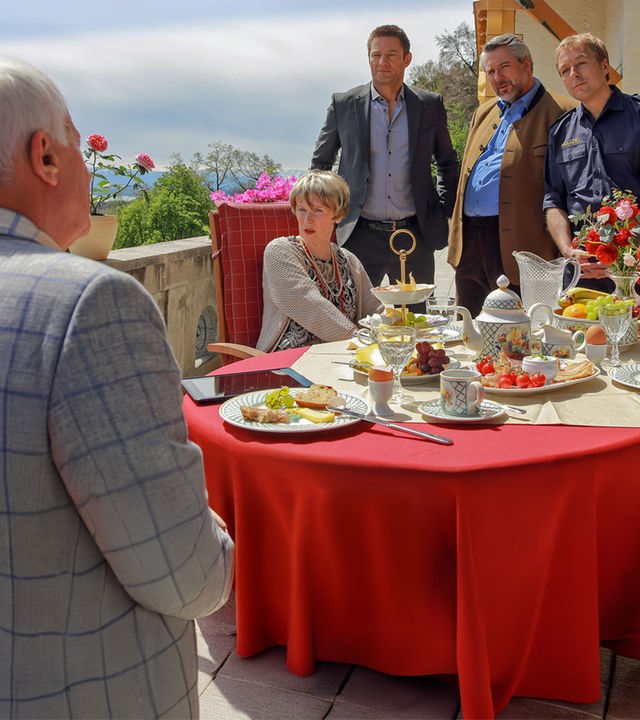 "Die Rosenheim-Cops - Ein ganz besonderes Event": Robert Pröbstl (Nikol Voigtländer) und Jutta Anzinger (Barbara de Koy) sitzen am gedeckten Frühstückstisch auf der Terrasse. Hinter Jutta Anzinger stehen die Rosenheim-Cops Hansen (Igor Jeftic), Stadler (Dieter Fischer) und Mohr (Max Müller), die Pröbstl ansehen.